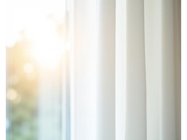Primer plano de una cortina blanca impecable recién limpiada y planchada, colgando elegantemente bajo la luz del sol que entra por una ventana, con un ligero pliegue que denota su suavidad y frescura. Refleja la calidad del servicio de lavado, planchado y reinstalación, resaltando la atención al detalle y el acabado profesional de Hilos Limpios.