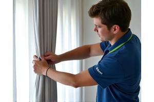 Un técnico de Hilos Limpios en Madrid, se concentra en descolgar cuidadosamente una cortina pesada de un riel, utilizando herramientas adecuadas y mostrando profesionalismo a la hora de manipular la tela, asegurándose de no dañarla ni el sistema de colgado y poniendo atención en el detalle para asegurar.